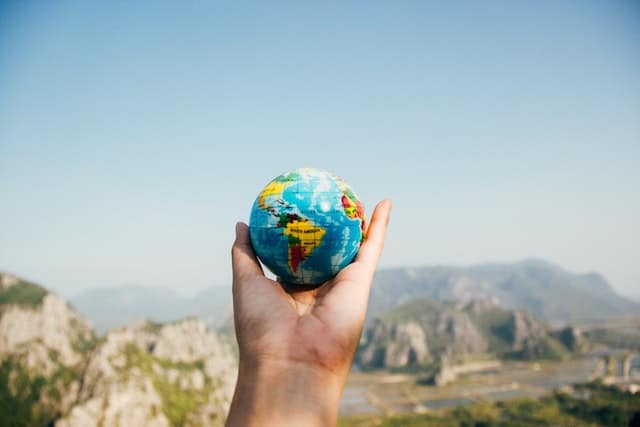 A person holding a world globe facing mountains.